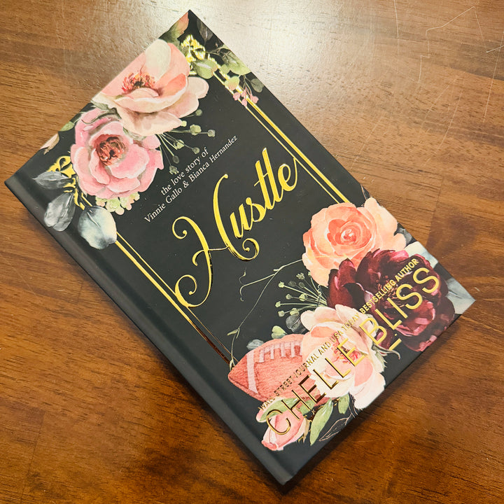 A hardcover book titled 'Hustle' by Chelle Bliss, featuring a black cover decorated with pink and red roses, green leaves, and gold accents, resting on a wooden table.