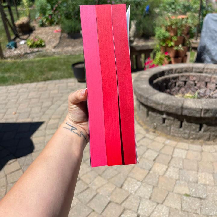 Hand holding three books, pages in red  in front of a garden background