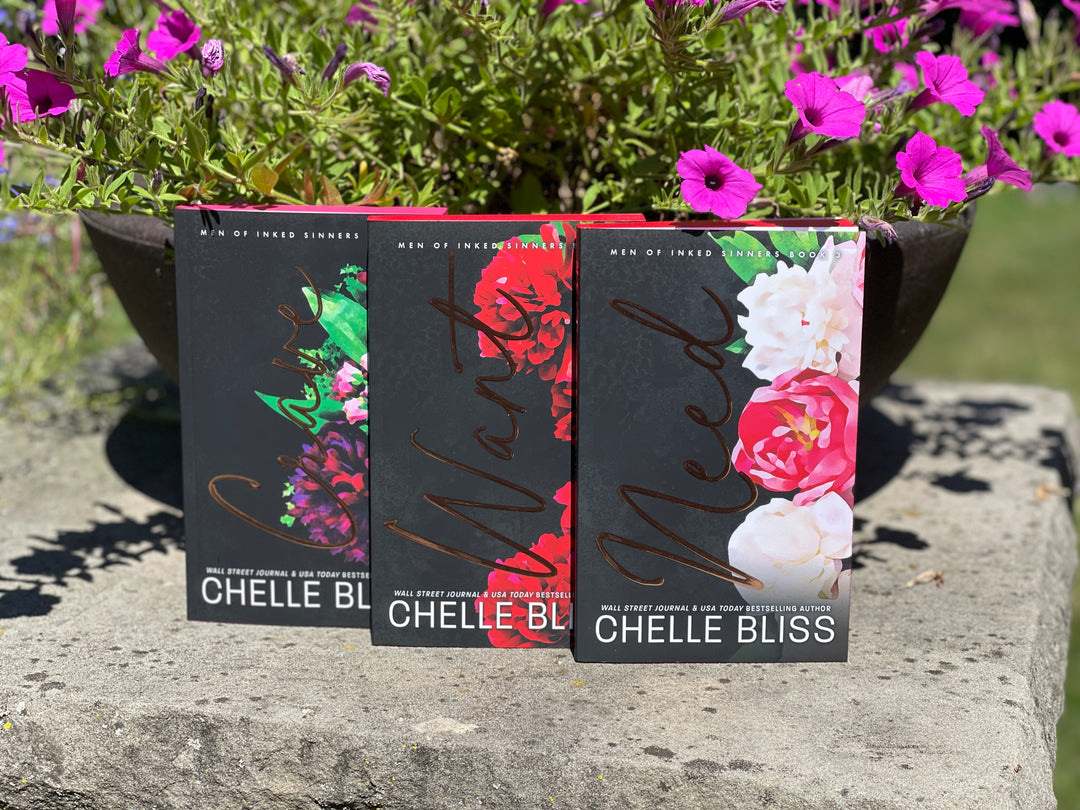 Three books by Chelle Bliss, Crave, Want and Need with floral designs on a stone surface with flowers in the background