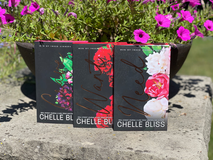 Three books by Chelle Bliss, Crave, Want and Need with floral designs on a stone surface with flowers in the background