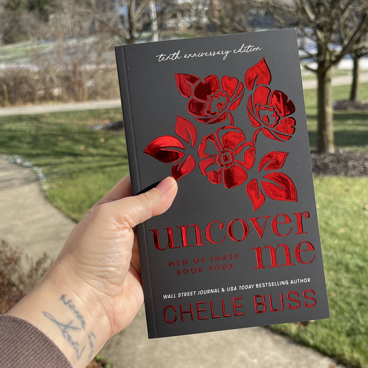 paperback book Uncover Me 10th Anniversary Foil Special Edition by chelle bliss with red flowers on the cover, held in a hand with grass in the background.
