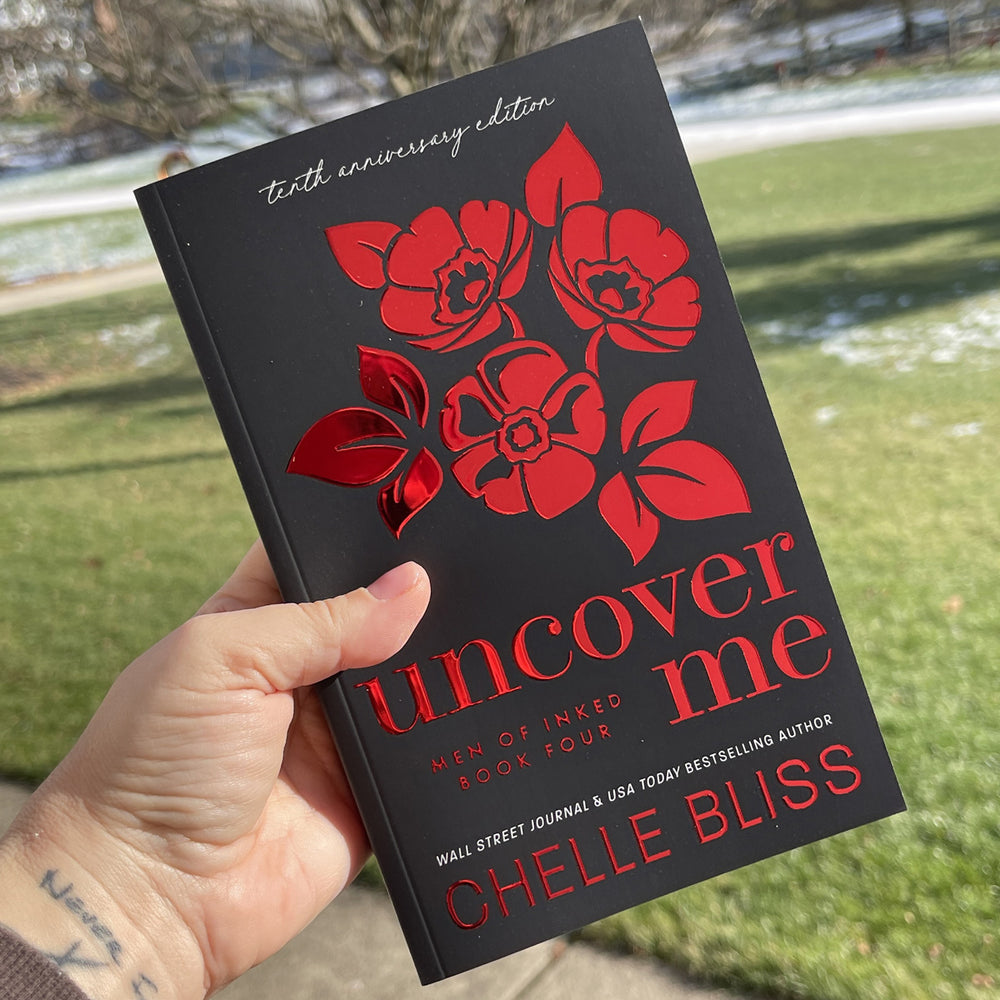 paperback book Uncover Me 10th Anniversary Foil Special Edition by Chelle Bliss featuring red flowers on the cover, held in a hand with grass in the background.